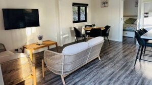 Una sala de estar moderna con un sofá, una mesa de comedor con sillas, un televisor montado en la pared y un ventanal con cortinas.