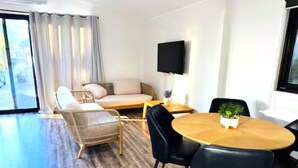 Una sala moderna con un sofá, una mesa de comedor con sillas, un televisor de pantalla plana y un gran ventanal con cortinas.