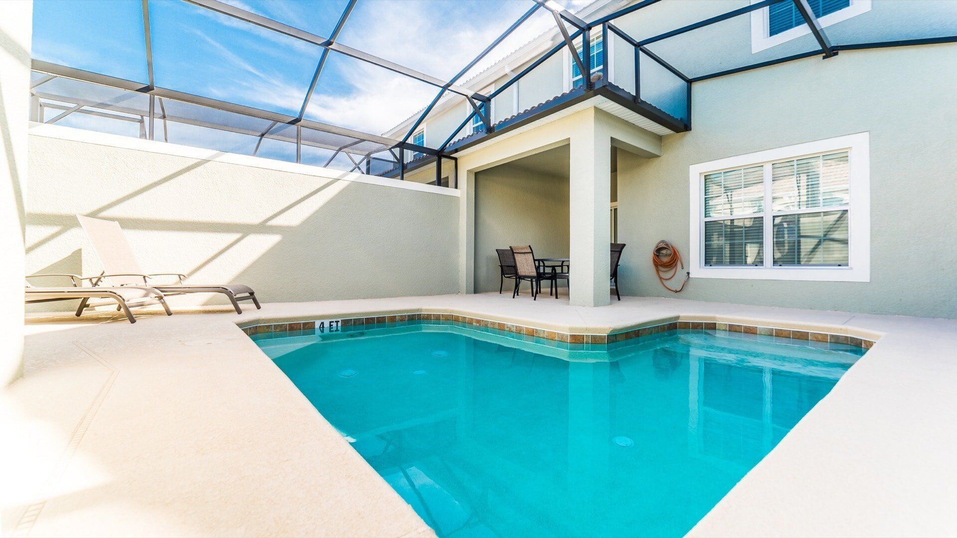 An indoor swimming pool with clear blue water, surrounded by a white tiled deck. There are lounge chairs and a table with chairs nearby.