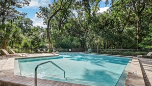 Una piscina rettangolare con bordo piastrellato, circondata da una recinzione metallica e ombreggiata da alberi.