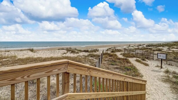 Ein hölzerner Steg führt zu einem Strand mit blauem Himmel und Wolken.