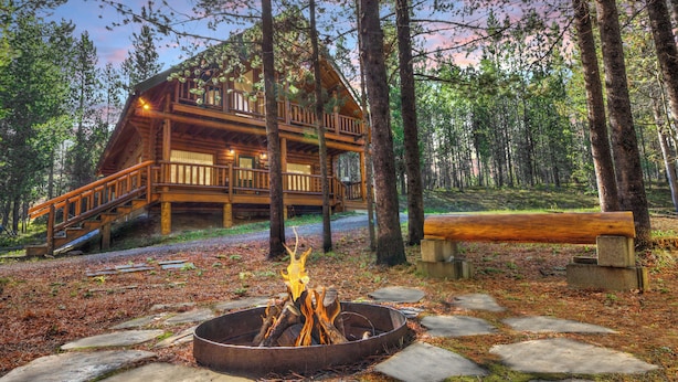 Uma cabana com varanda, lareira e um banco em meio a uma floresta.