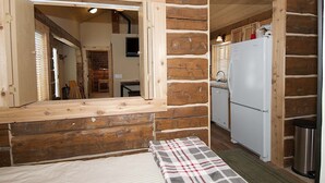 A log cabin interior with a wooden-framed window, a kitchenette with a white refrigerator, and a small bed with a plaid blanket. - True Grit Adventure (Island Park)