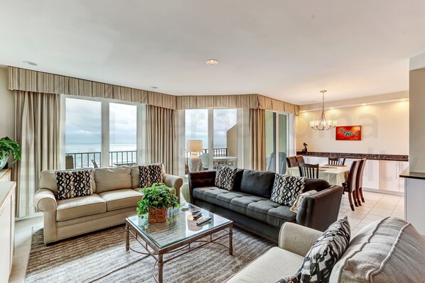 A spacious living room with a sofa, armchair, and a coffee table. There is a dining area with a table and chairs, and a balcony with curtains. - 1876 Turtle Dunes Amelia Island Plantation (Fernandina Beach)