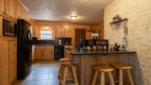 A kitchen with wooden cabinets, a black refrigerator, a sink, and a window. There are stools at the counter. - Remington Reload (Island Park)
