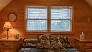 A cozy room with a large window, a bed with patterned bedding, a nightstand with a lamp, and a small table with a tissue box. - Remington Reload (Island Park)