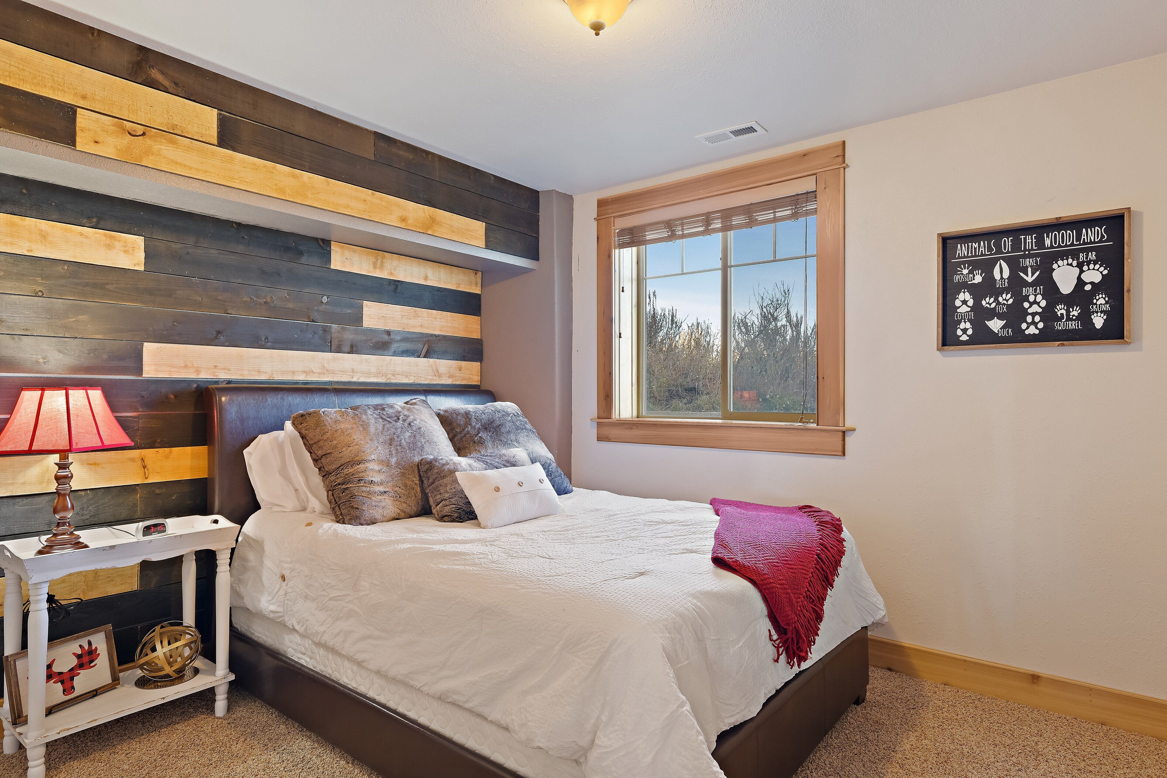 Une chambre à coucher comprenant un lit, une table de chevet, une lampe, un panneau décoratif fixé au mur et une fenêtre donnant sur des arbres.