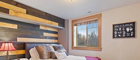 A bedroom with a bed, a nightstand, a lamp, a wall-mounted sign, and a window with a view of trees.