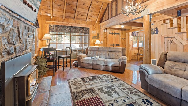 A cozy cabin interior with a stone fireplace, a plush sofa, a dining area, and a staircase leading to an upper level. - Double Loop (Island Park)