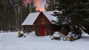 A wooden cabin with a red door, surrounded by snow-covered ground and trees. - Double Loop (Island Park)