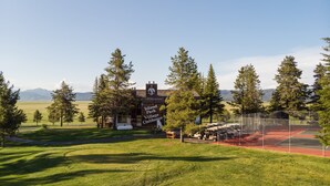 Una club house con un cartello che riporta "Island Park Village Clubhouse", circondata da un campo da tennis recintato e un prato alberato.