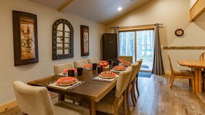 Un coin repas avec une longue table dressée pour six personnes, des chaises en bois, un ventilateur de plafond et une fenêtre avec des rideaux.