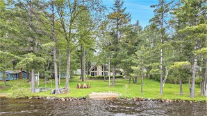 A lakeside property with a house surrounded by trees, a grassy area with outdoor furniture, and a small stream. - Relax & Explore Waterfront Crawford Lake Cottage 4878 (Kalkaska)