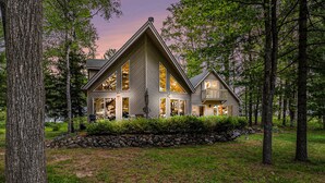 A large, modern house with a prominent triangular roof, surrounded by a stone wall and lush greenery. - Relax & Explore Waterfront Crawford Lake Cottage 4878 (Kalkaska)