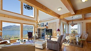 A spacious living room with wooden beams, a stone fireplace, a television, and a view of a lake and mountains. - Henrys Lake Goose Bay (Island Park)