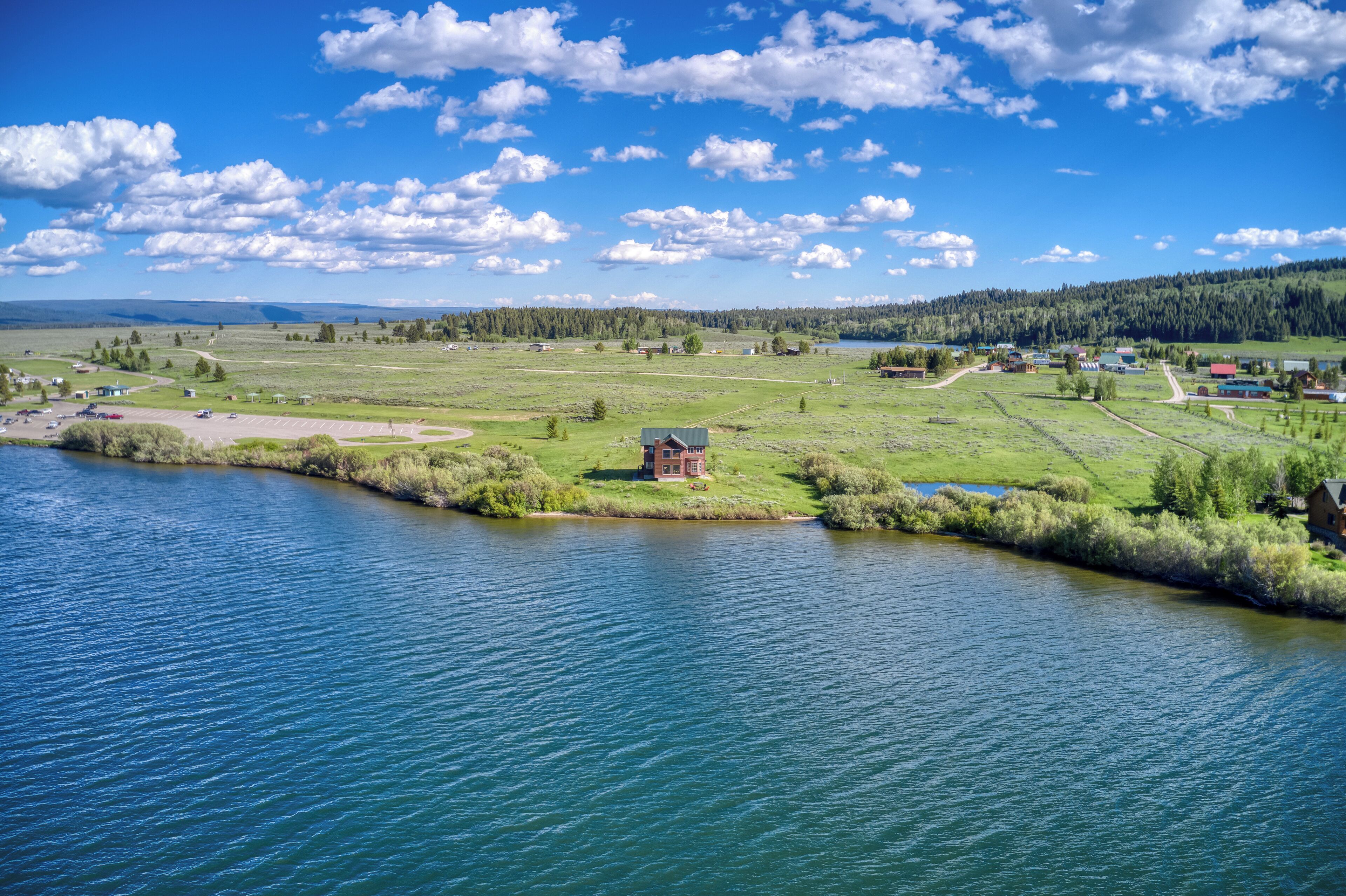 A lakeside view with a small building near the water, surrounded by open fields and a forested area in the background.