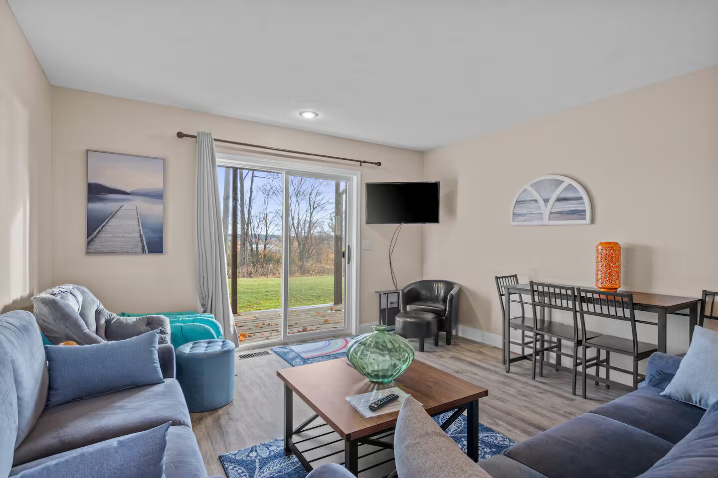 A spacious living room with a sofa, armchair, and a dining table with chairs. A TV is mounted on the wall, and there is a large window with a view of the outdoors.