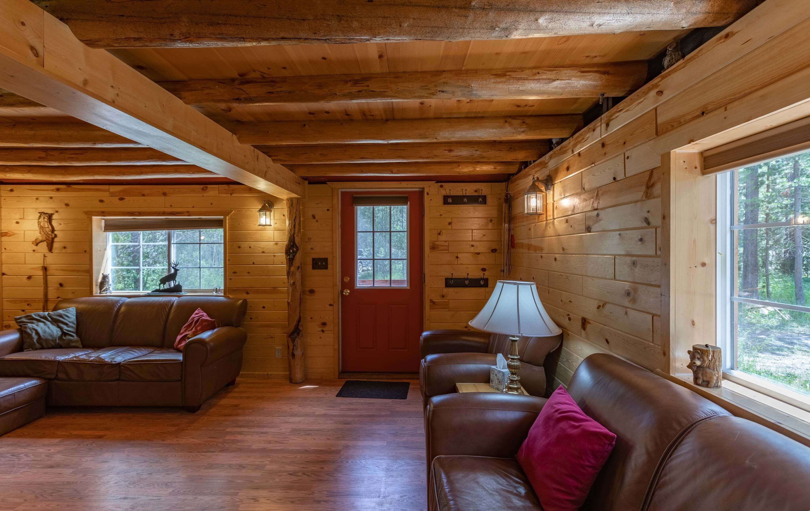 L’intérieur d’une cabane avec des murs en bois, une porte rouge, deux canapés en cuir, une lampe de table et une fenêtre donnant sur la forêt.