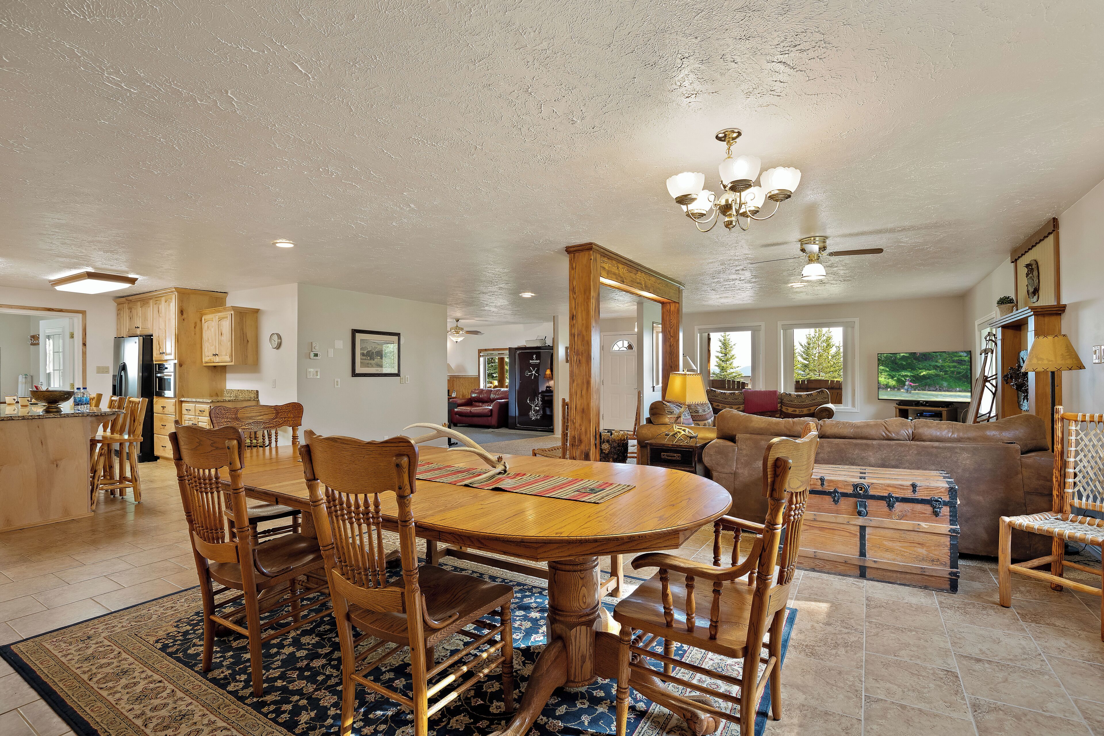 A spacious living area with a wooden dining table, chairs, and a large sofa. There is a kitchen in the background with wooden cabinets and a refrigerator.