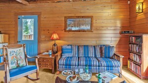 Ein gemütliches Cabin-Interieur mit Holzbalkendecke, einer blauen Tür, einem gestreiften Sofa, einem Schaukelstuhl, einem Bücherregal und einem Esstisch mit Tellern und einer Schüssel.
