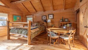 Ein Holzblockhaus-Interieur mit Bett, Esstisch, Stühlen, einer Lampe und gerahmten Bildern an den Wänden.