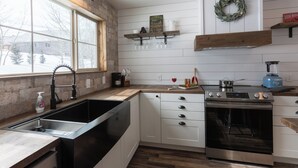 A kitchen with a large window, white cabinetry, a stainless steel sink, and an oven. - Cast Iron Moose (Island Park)