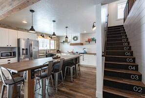 Una cocina moderna con una isla central, taburetes de bar, electrodomésticos de acero inoxidable y una escalera de madera con escalones numerados.