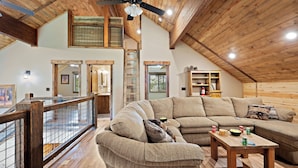 A spacious loft with a high ceiling, wooden beams, a large sectional sofa, a wooden coffee table, and a view through a glass railing. - Set the Hook (Island Park)