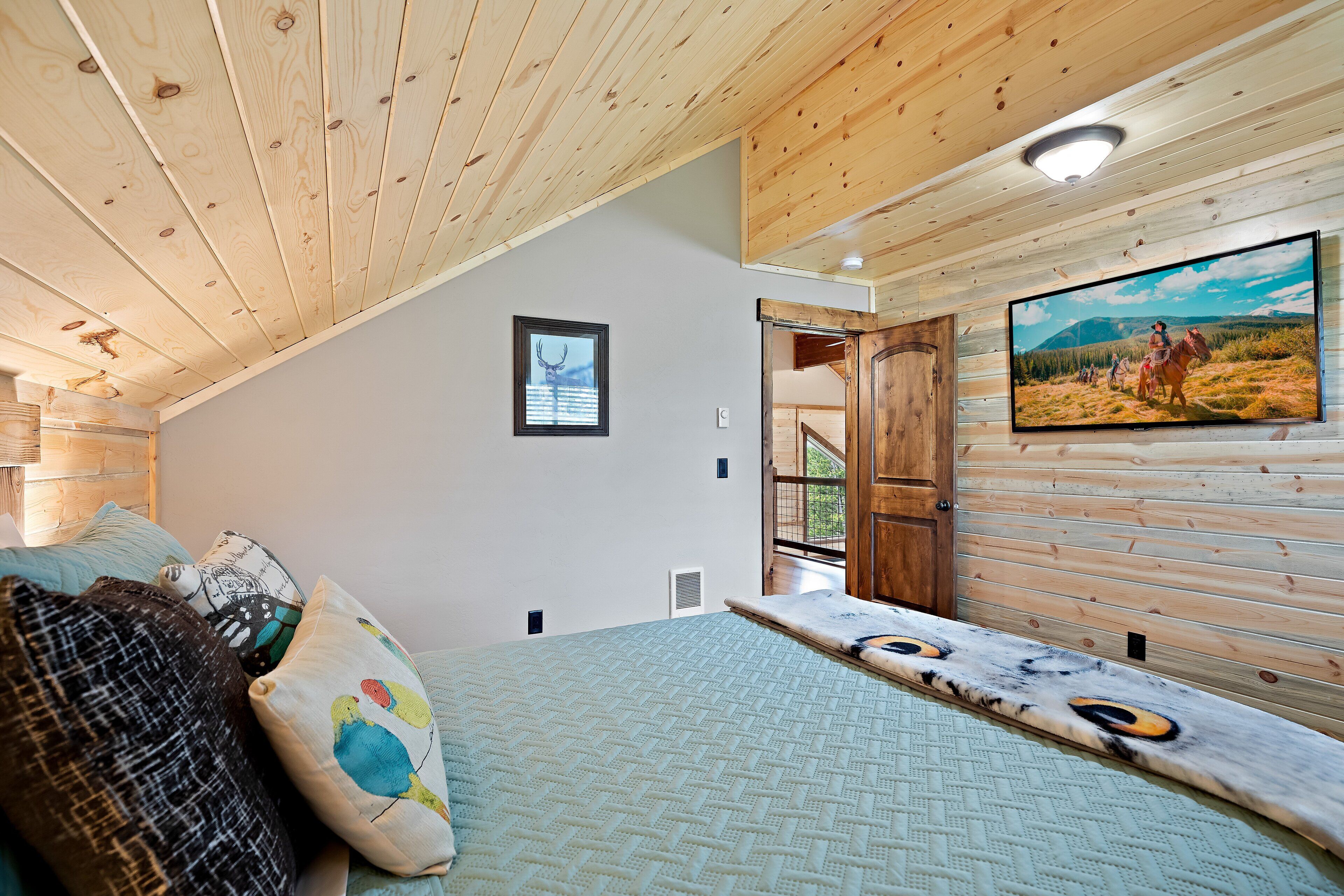 A bedroom with a sloped ceiling, a bed with patterned bedding, a mounted TV, and a framed picture on the wall.