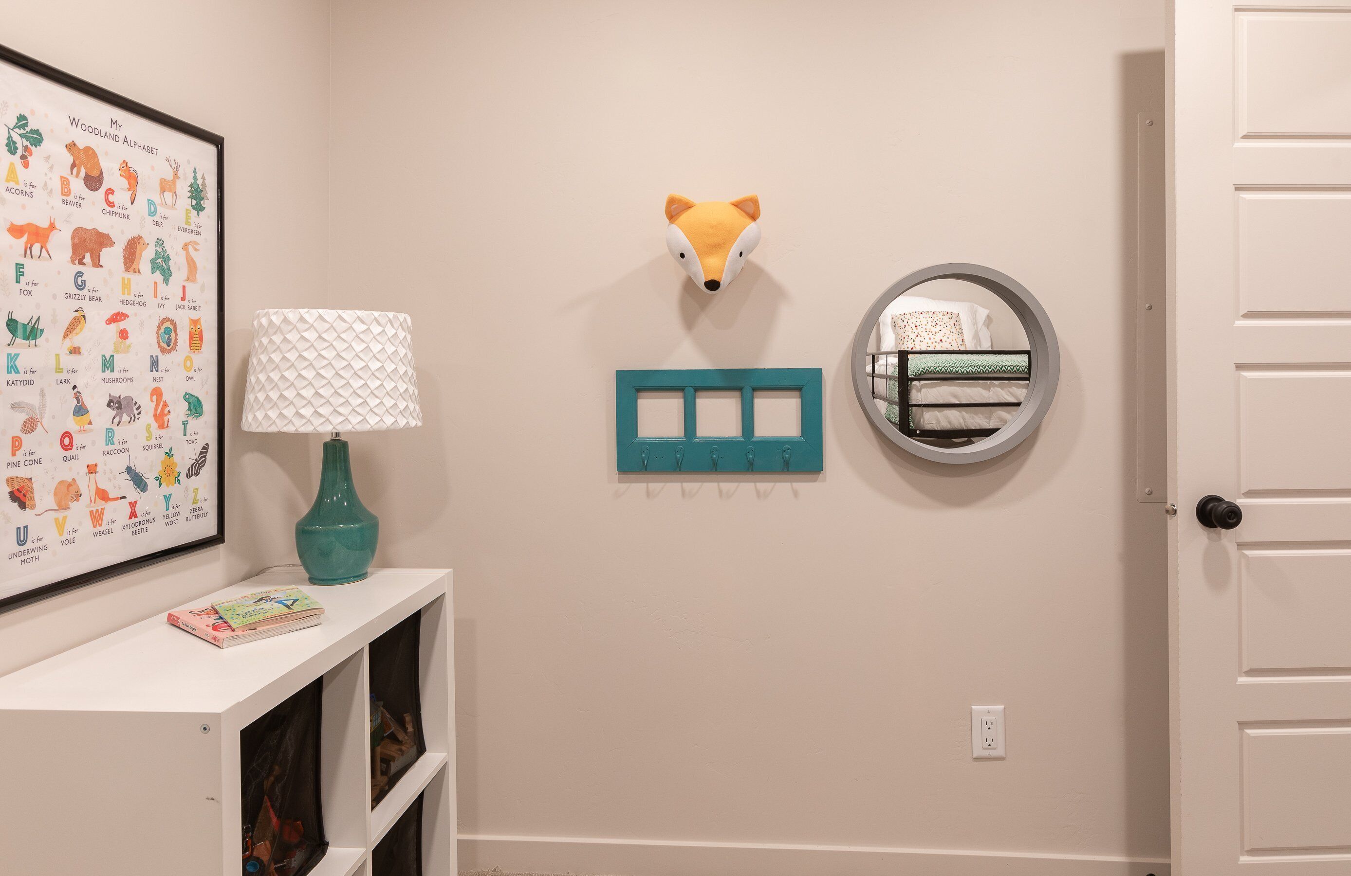 A hallway with a framed alphabet poster, a teal lamp, a fox head decoration, a circular mirror, and a teal coat rack.
