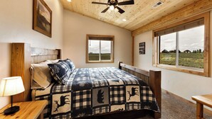 Ein Schlafzimmer mit Holzboden, einem Bett mit kariertem Überwurf, einem Nachttisch, einem Fenster mit Blick ins Freie und einem Deckenventilator.