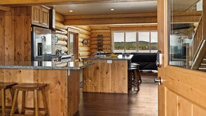 Ein Holzblockhaus-Interieur mit einer Küche, die einen Edelstahl-Kühlschrank, Holzschränke und ein großes Fenster mit Blick auf eine bewaldete Landschaft zeigt.