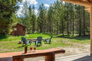 A wooden deck with a table set for two, overlooking a forested area with a small cabin in the background. - Little Fox (Island Park)