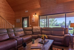 A wooden cabin interior with a leather sectional sofa, a coffee table with a book and a vase of flowers, and a large window offering a view of the outdoors. - Deer View (Island Park)