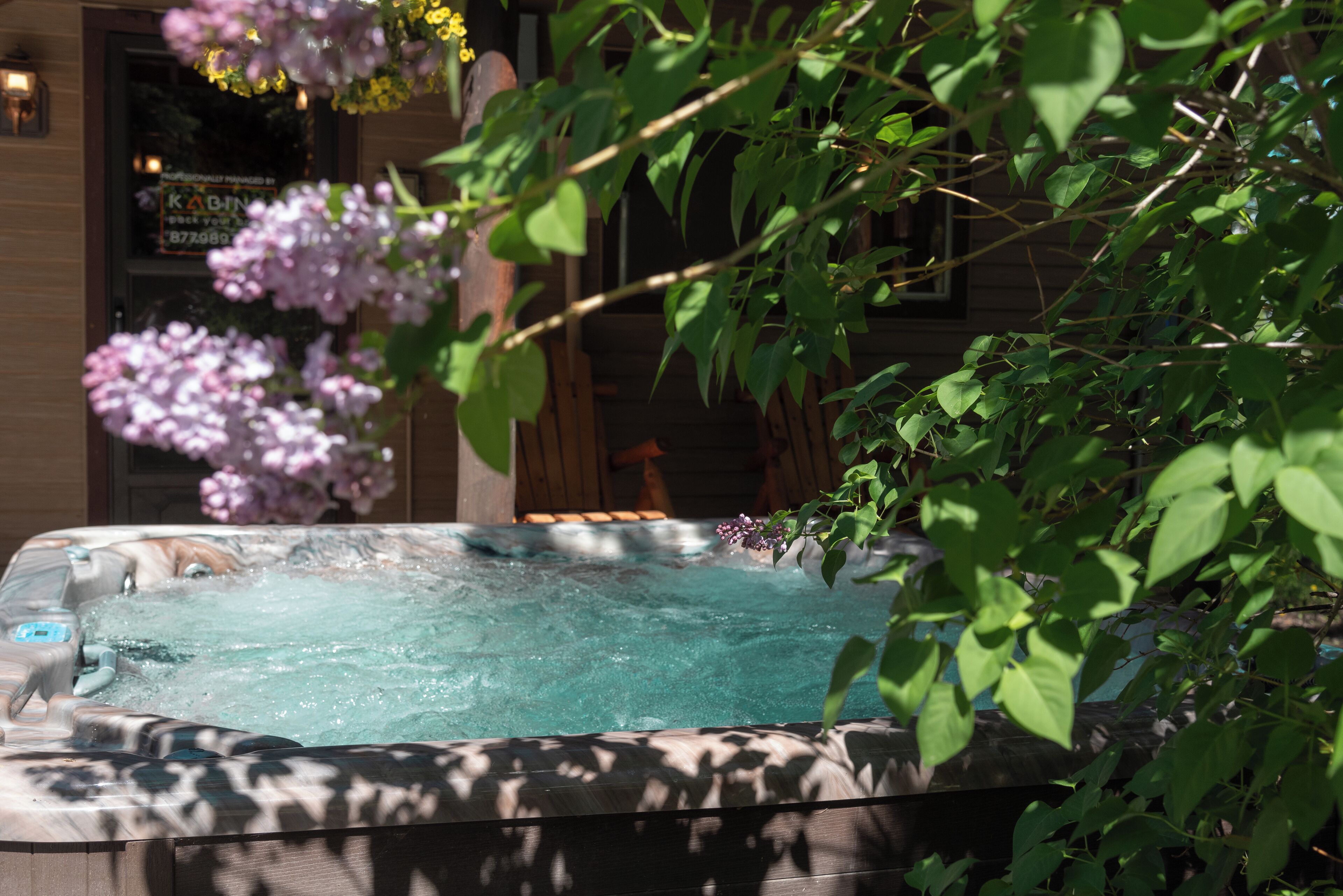 A hot tub with a wooden deck, surrounded by greenery and a building with a sign.