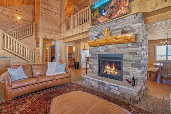 A spacious living room with a stone fireplace, leather sofa, and a high ceiling with wooden beams. - Liberty Lodge (Island Park)