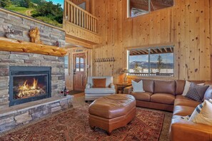 A cozy living room with a stone fireplace, leather sofa, and a view of a snowy mountain. - Liberty Lodge (Island Park)