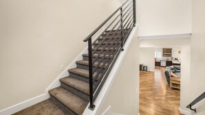 Eine Treppe mit Teppichstufen und einem Metallgeländer, die zu einem Wohnbereich mit Sofa und einer dahinterliegenden Küche führt.