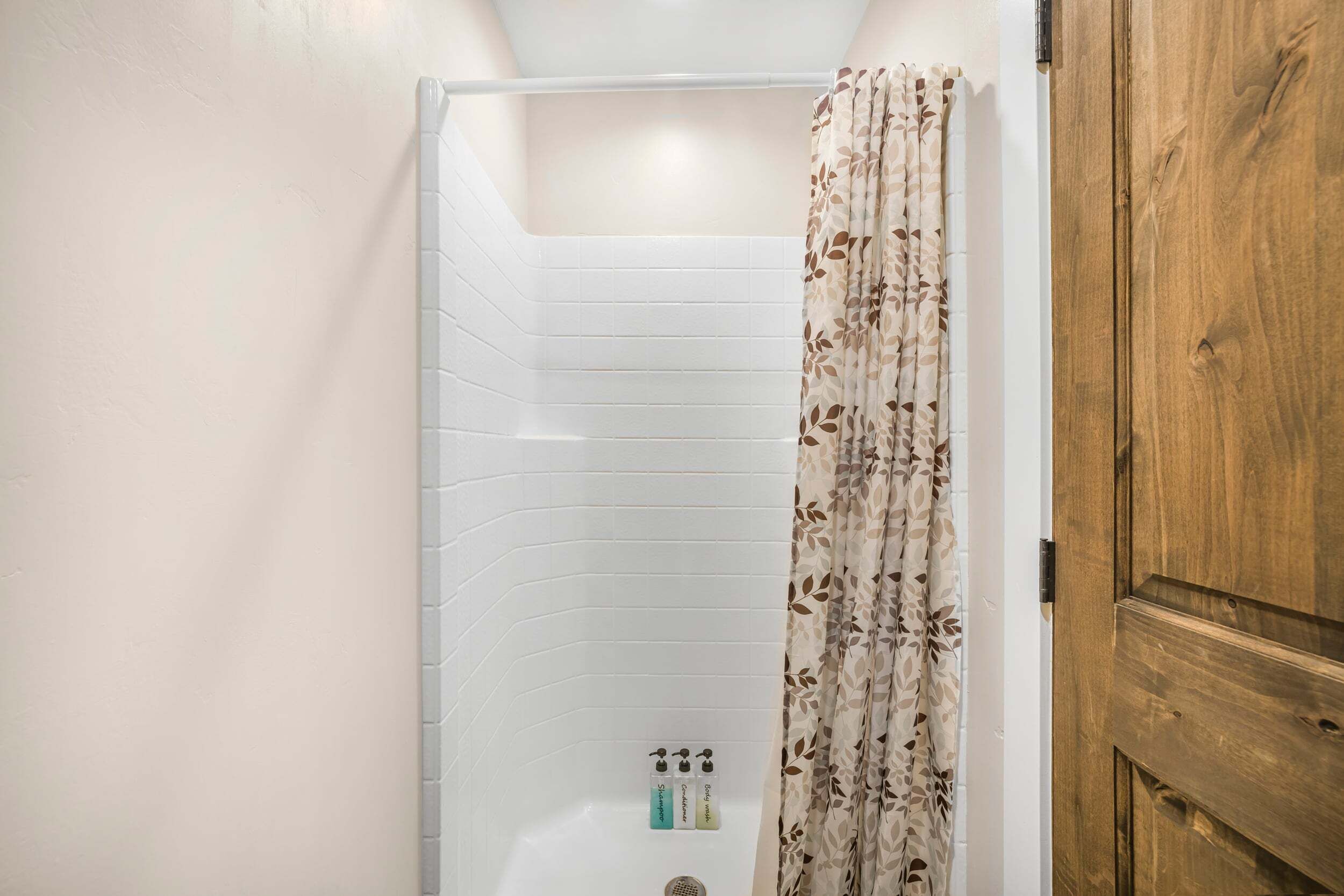 A shower with a white tiled wall, a wooden door, and a floral shower curtain.