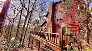 A rustic log cabin with a wooden deck and a chimney, surrounded by trees.