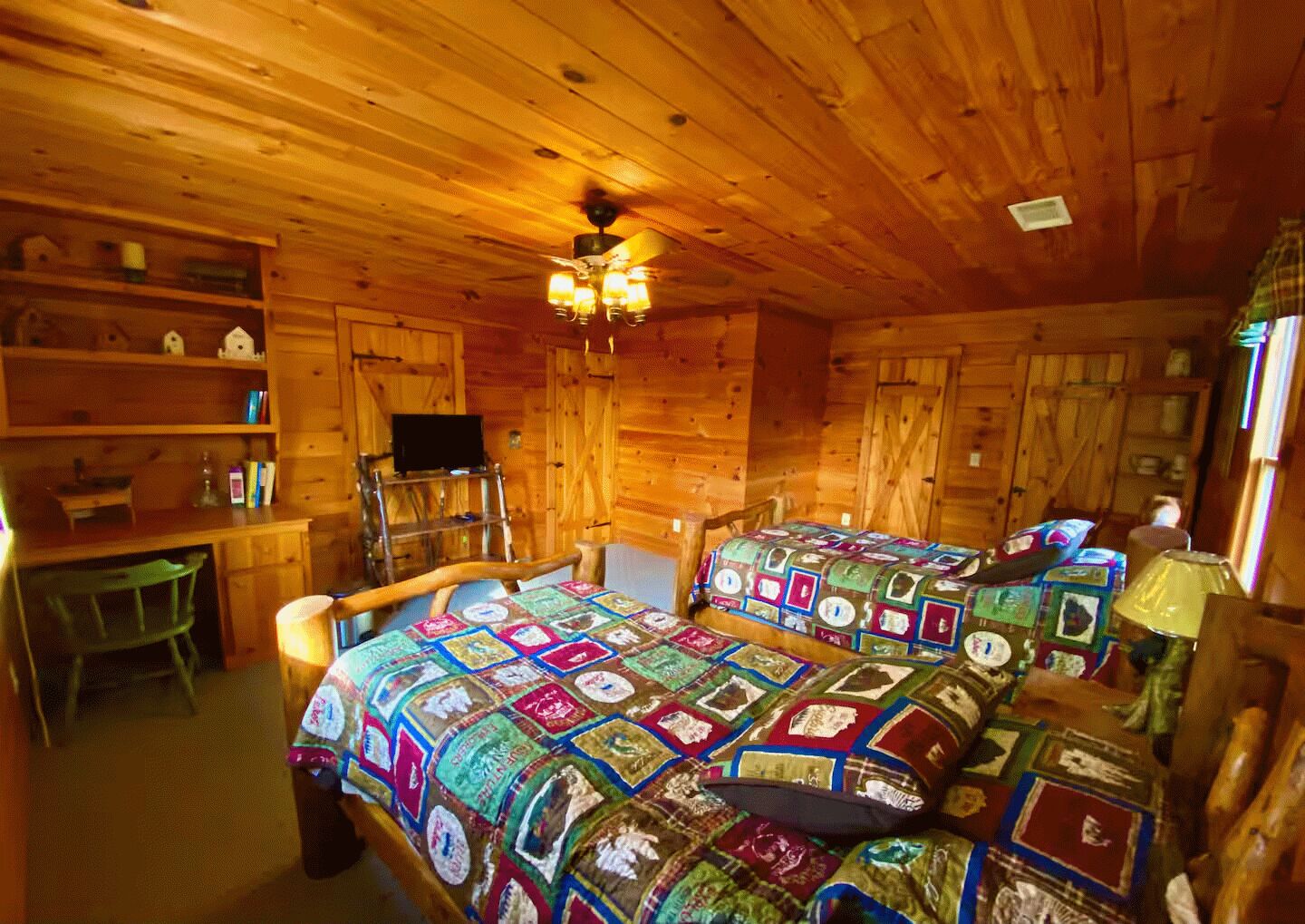 A cozy cabin room with wooden walls and ceiling, two beds with colorful quilts, a desk with a chair, and a television on a shelf.