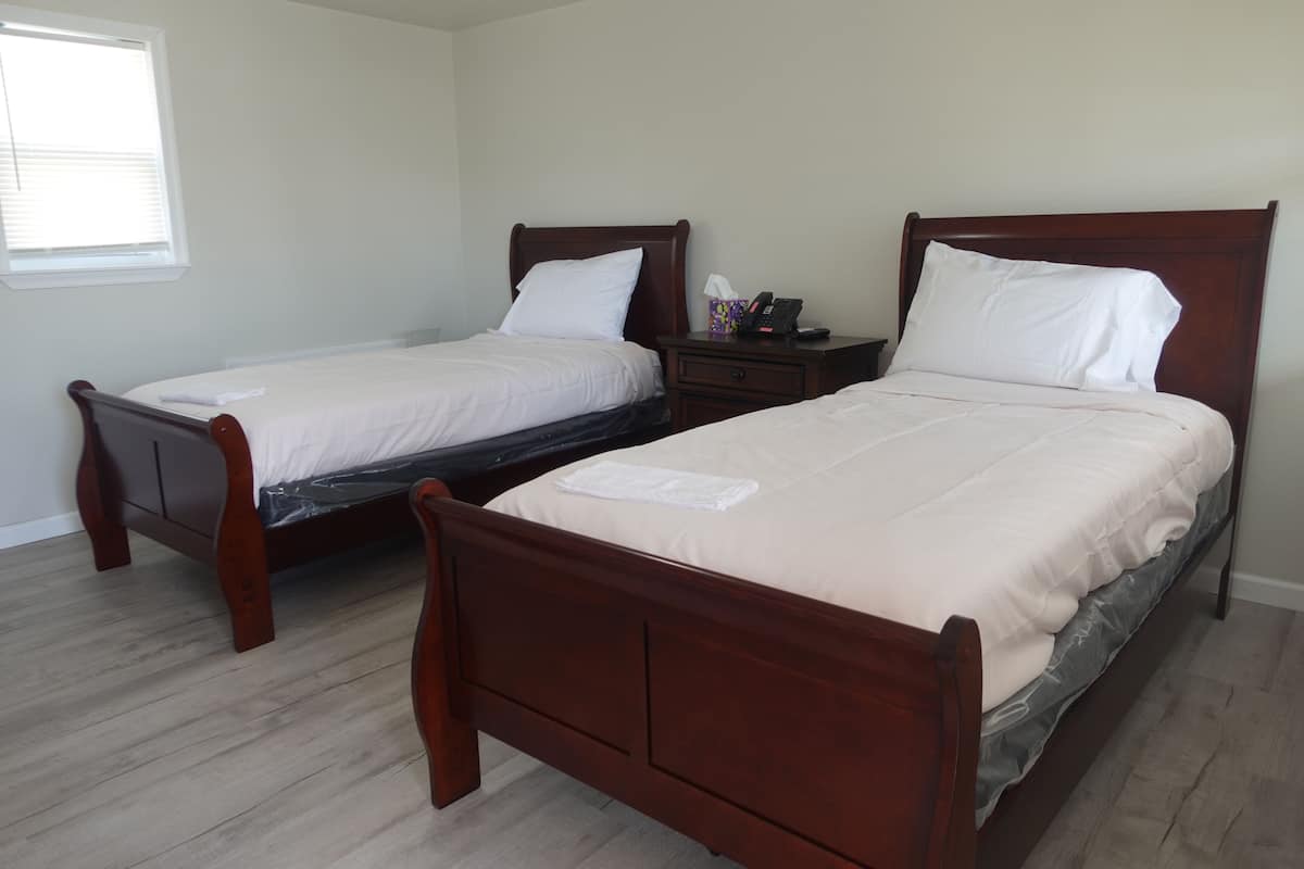 A hotel room with two wooden beds, a nightstand with a phone and tissues, and a window with blinds.