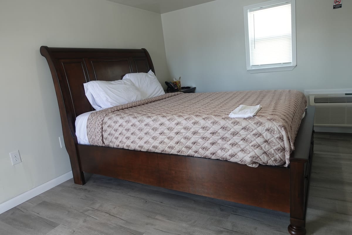 A neatly made bed with a patterned bedspread and pillows, a wooden headboard, a bedside table with a phone, and a window with blinds.