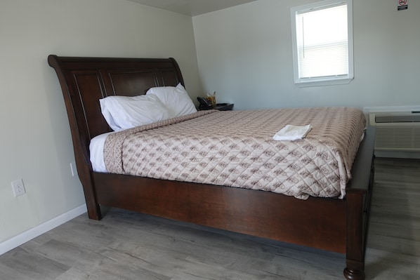 A neatly made bed with a patterned bedspread and pillows, a wooden headboard, a bedside table with a phone, and a window with blinds. - Freedom Inn Motel (Grantville)