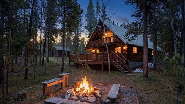 Una cabaña con terraza, un pozo de fuego y un bosque circundante.