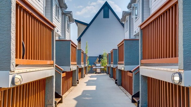 Una hilera de casas modernas de un piso, con cercas de madera y un camino central que conduce a un edificio grande de color blanco con techo azul.