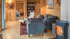Een gezellige woonkamer met een bank, fauteuil en een open haard. Er staat een boekenkast en een lamp in de kamer.