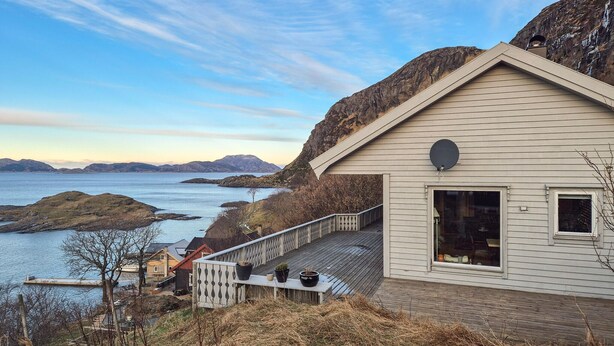 Een huis aan de kust met een terras dat uitkijkt over een waterpartij en heuvels.