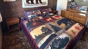 Une chambre avec un plafond en bois, un ventilateur de plafond, un lit recouvert d’une courtepointe ornée de motifs représentant la forêt et la faune, une table de chevet, une commode et un miroir fixé au mur.