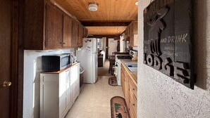 Une cuisine étroite avec des meubles en bois et un réfrigérateur blanc. Un tableau noir accroché au mur annonce les plats et les boissons proposés.